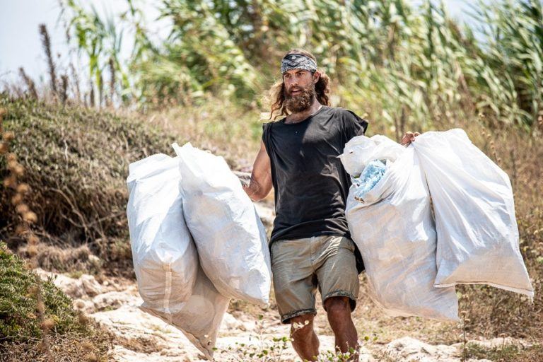 a man carrying two bags of garbage on a dirt road.