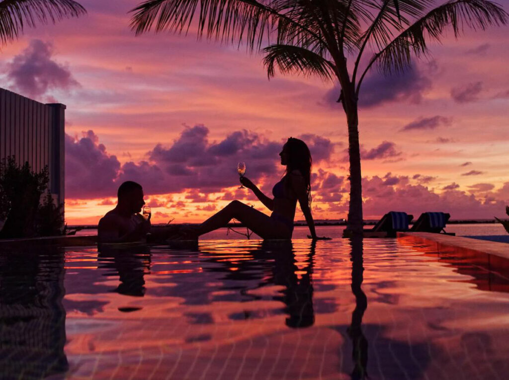 a couple sitting on the edge of a swimming pool at sunset