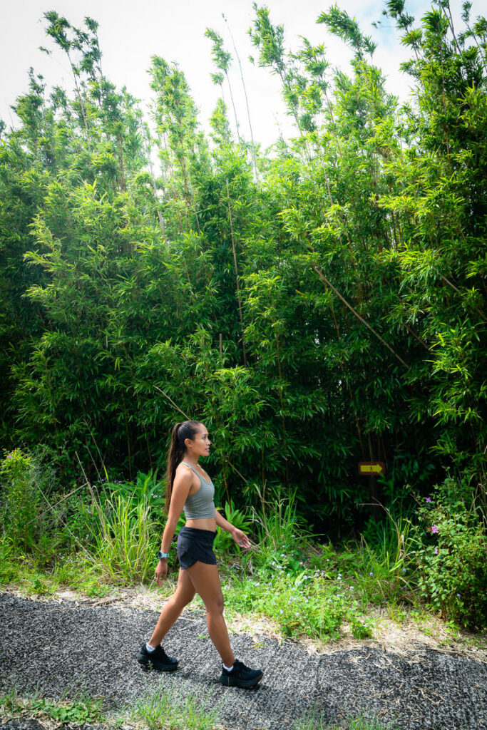 a woman walking down a dirt road next to a lush green forest.