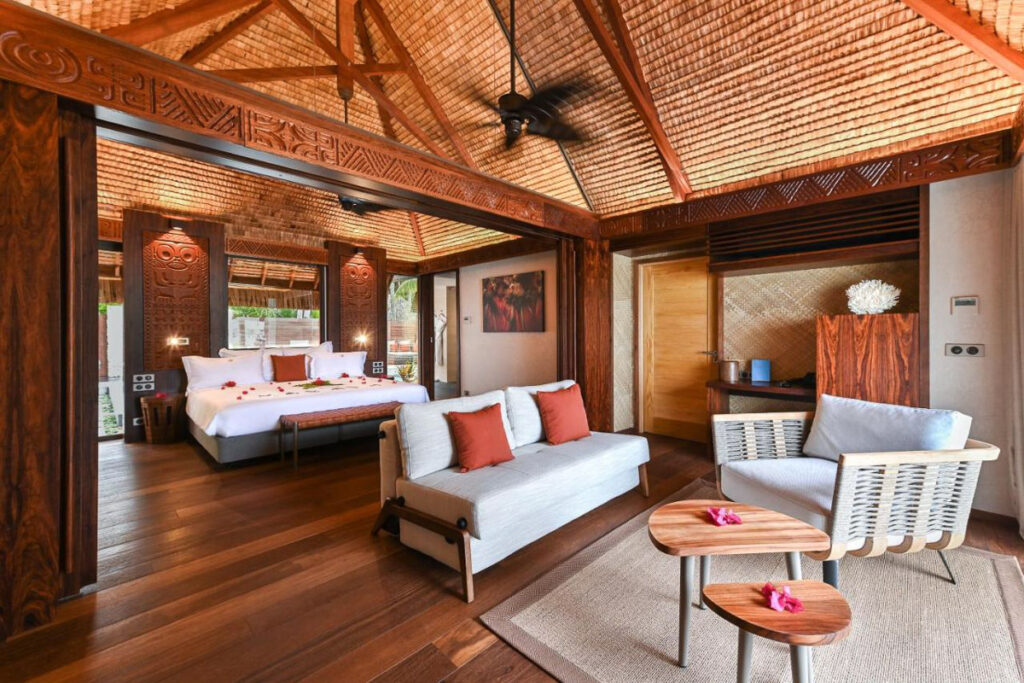 An overwater bungalow in French Polynesia with a bed, couch, chairs and a ceiling fan.