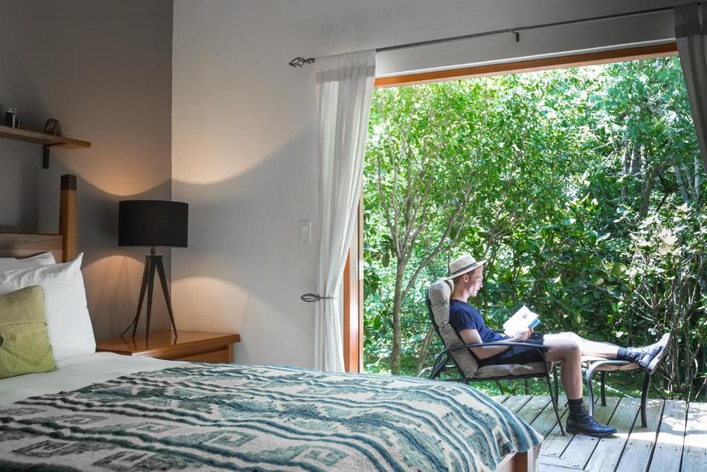 A man reading a book in a bedroom with a view of the trees.