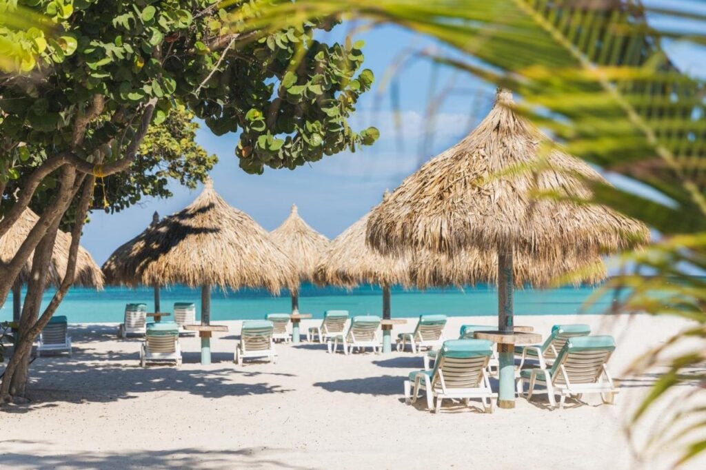 Beach scene with several thatched umbrellas and lounge chairs arranged in the sand near the turquoise water, surrounded by lush greenery.