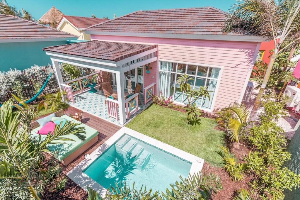 A pink house with a covered patio, lounge furniture, a small pool, and a well-maintained yard with various plants and trees.