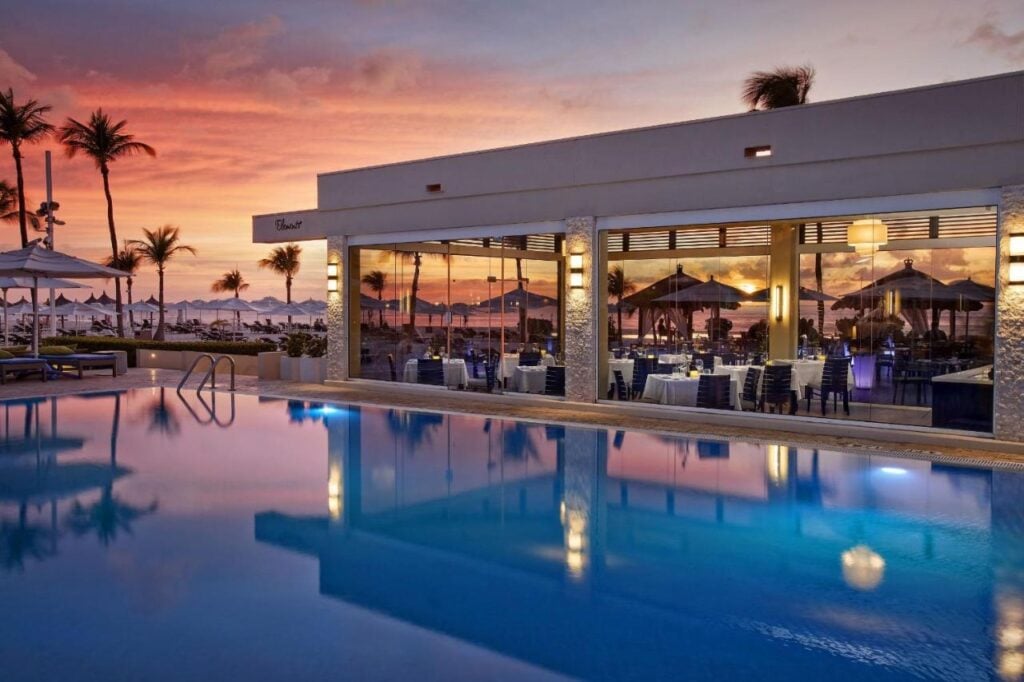 A glass-walled restaurant reflecting sunset colors is adjacent to a calm swimming pool, with outdoor seating under umbrellas and palm trees in the background.