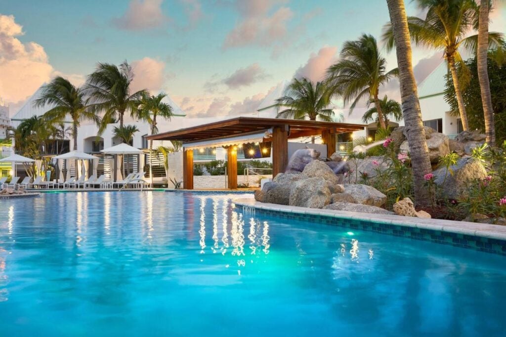 A serene pool area at sunset in one of Aruba's adults-only all-inclusive resorts, with palm trees, lounge chairs, cabanas, and rock formations surrounding the pool. The sky is partly cloudy, and lights are illuminating the pergola and adjacent area.