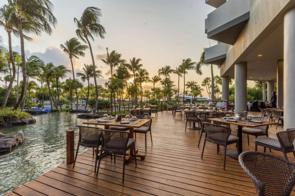 Outdoor restaurant with wooden deck overlooking a serene lagoon surrounded by palm trees, set against a sunset sky. Tables and chairs are arranged for dining, offering an idyllic experience typical of adults-only all-inclusive resorts in Aruba.