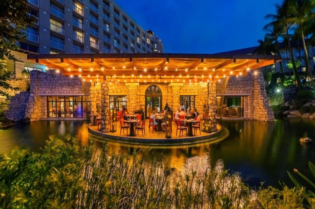 Outdoor restaurant with stone facade and string lights, surrounded by water, located next to a multi-story building at dusk. Diners are seated at tables under the lit pergola, reminiscent of the intimate settings found in adults-only all-inclusive resorts in Aruba.