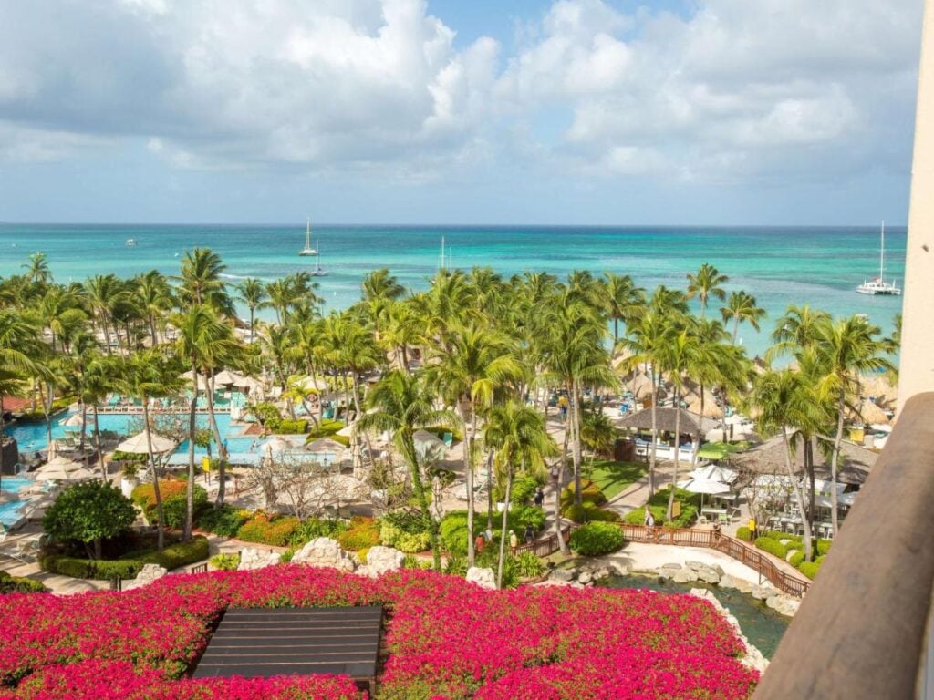 A tropical adults-only all-inclusive resort with lush palm trees, vibrant flowers, swimming pools, and a view of the turquoise ocean under a cloudy sky. Sailboats are visible in the distance, providing an enchanting backdrop for this paradise in Aruba.