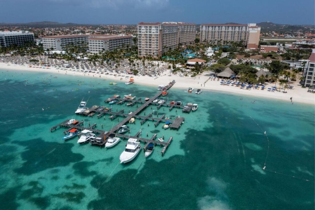 Aerial view of an adults-only all-inclusive beach resort in Aruba with multiple buildings, numerous beachgoers, and boats docked at a pier extending into turquoise waters.
