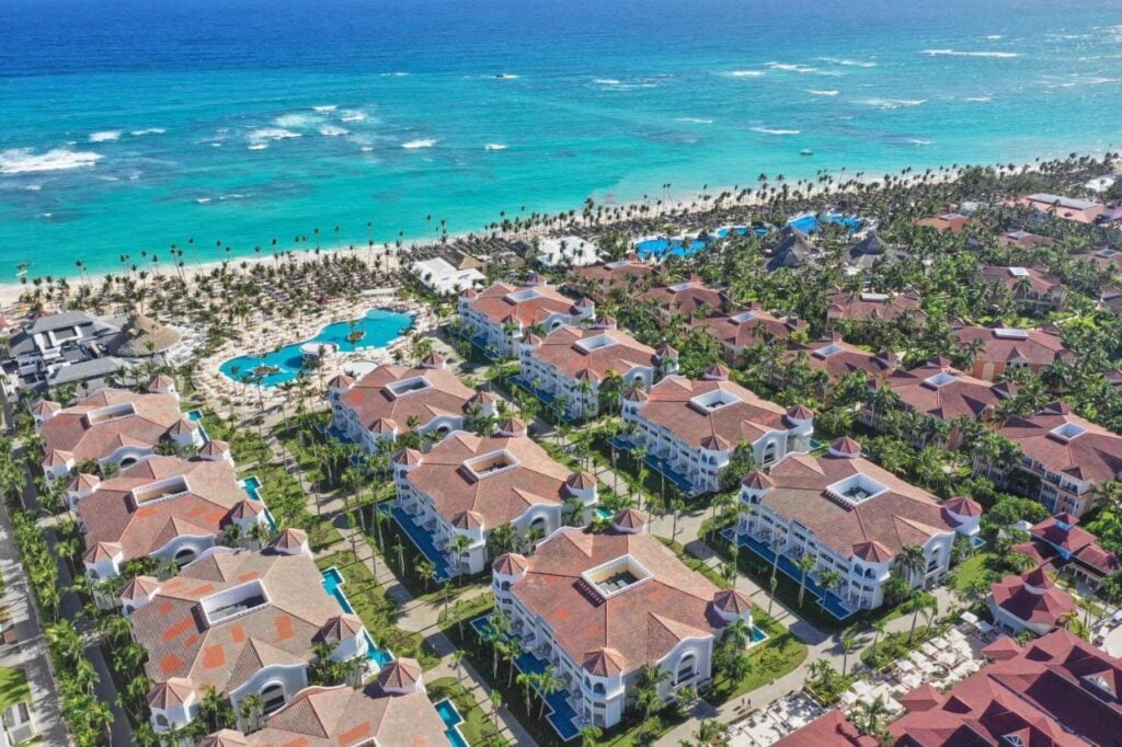 Aerial view of a seaside resort featuring numerous buildings with red roofs, multiple swimming pools, and lush green palm trees, adjacent to a beach with clear blue ocean water.