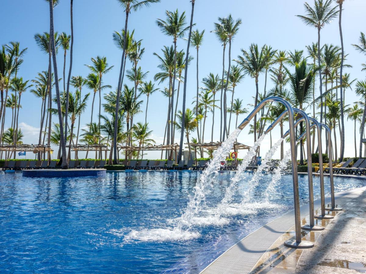An outdoor pool with several water fountains and lined with palm trees under a clear blue sky. Lounge chairs and shaded cabanas are visible in the background.