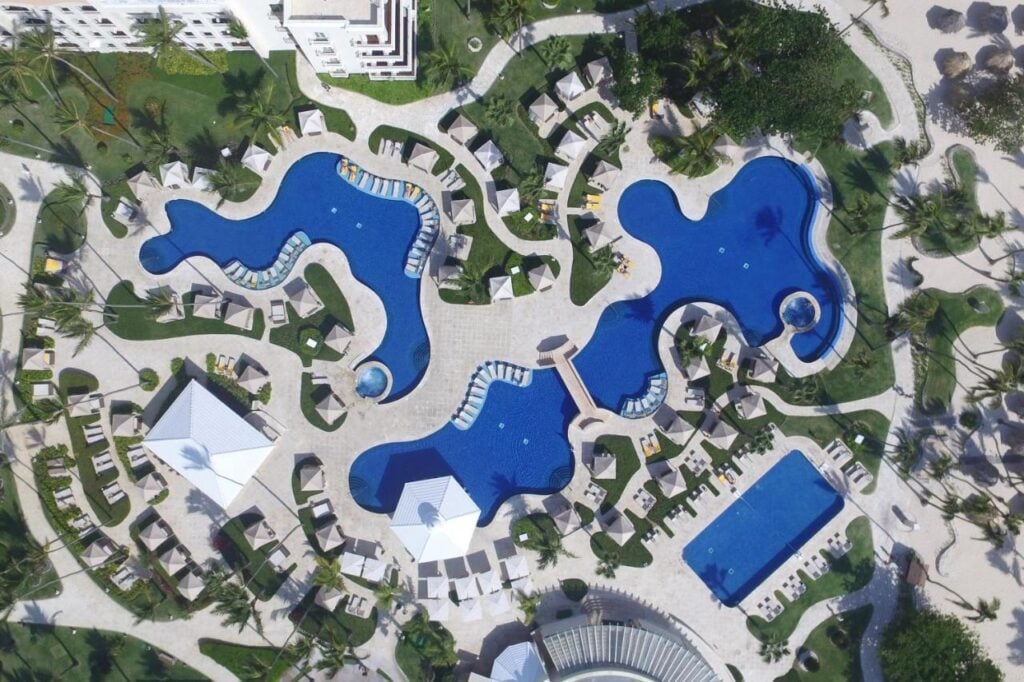 Aerial view of a resort with multiple irregularly shaped blue swimming pools surrounded by lounge chairs, green lawns, and palm trees. Several buildings and pathways are visible in the background.