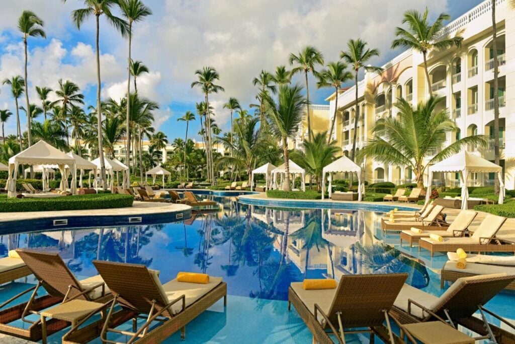 Lounge chairs and cabanas surround a large outdoor pool at a resort with palm trees and a multi-story building in the background under a partly cloudy sky.