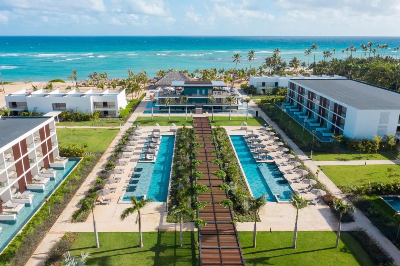 Aerial view of a beachfront resort with two large swimming pools flanked by palm trees, modern buildings, and the ocean in the background.