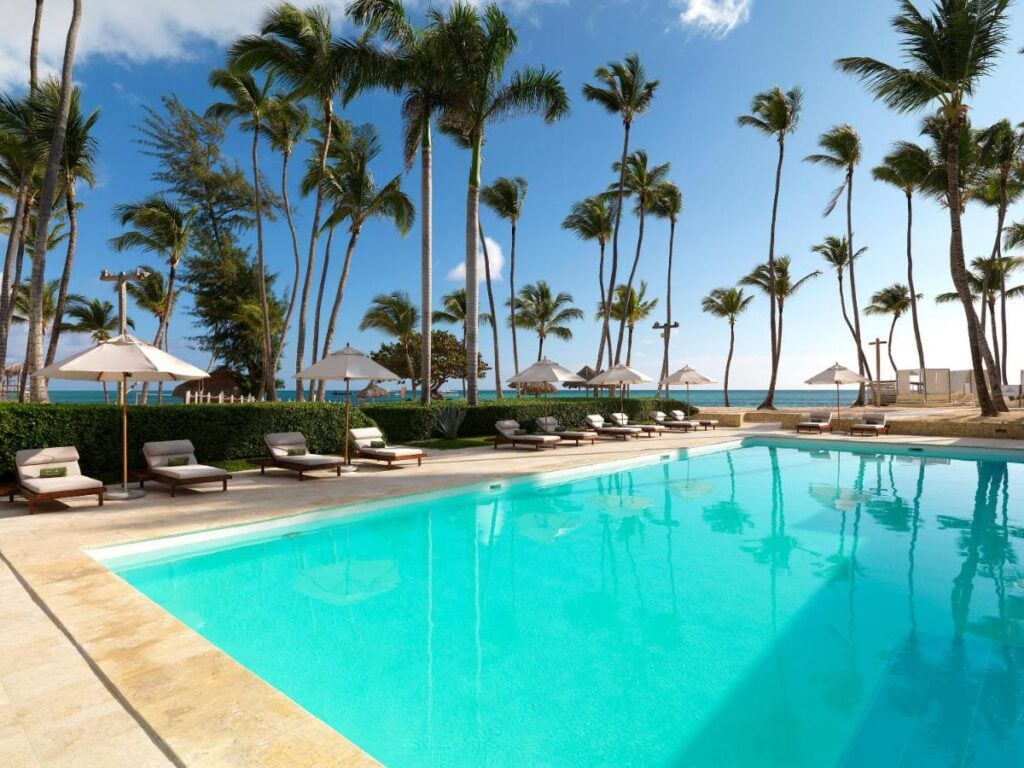 Outdoor swimming pool with lounge chairs, umbrellas, and tall palm trees around, set against a backdrop of a clear blue sky and ocean.