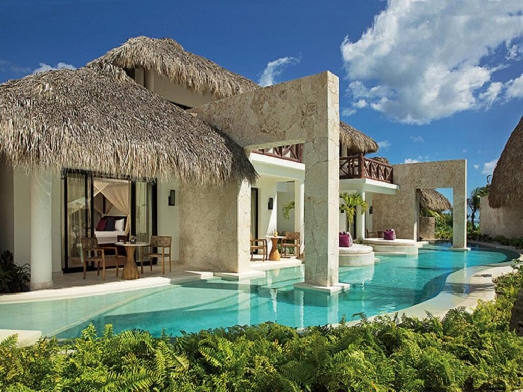 A luxurious villa with a thatched roof features modern stone architecture and a curved swimming pool surrounded by lush greenery.