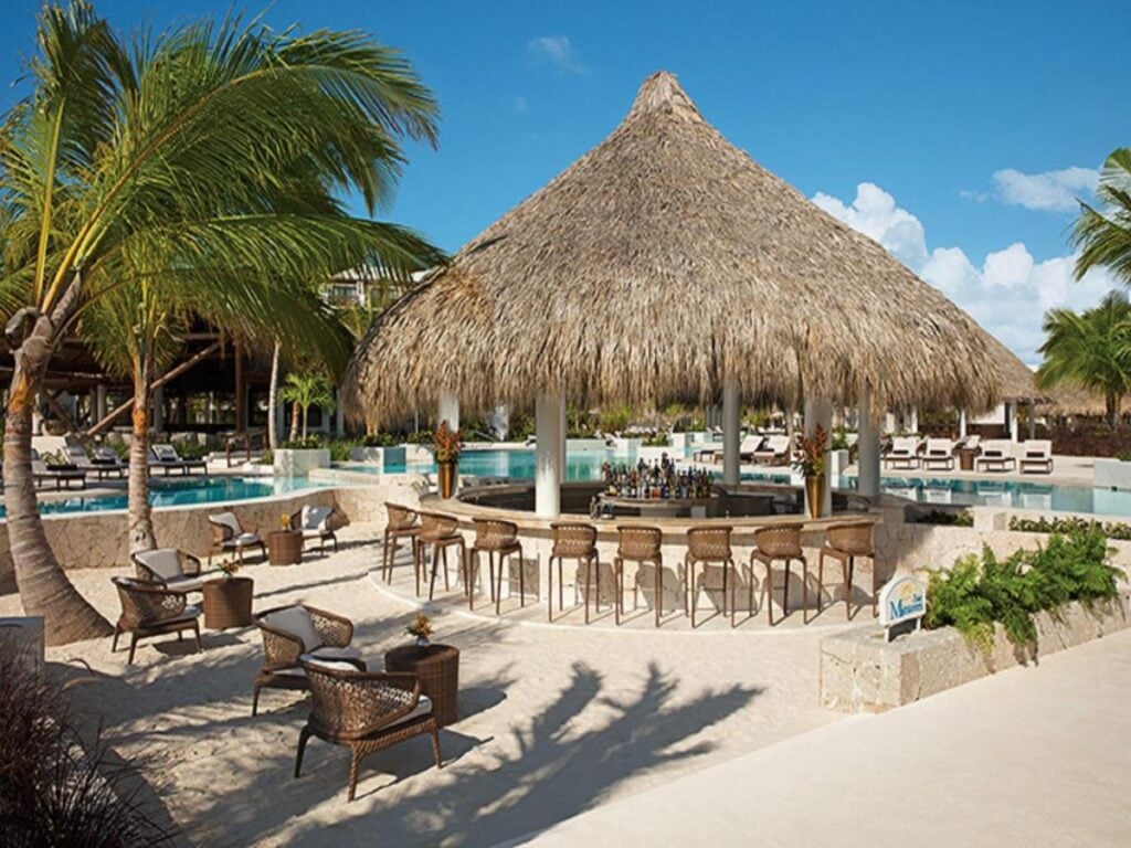 Outdoor tropical bar with a thatched roof surrounded by wicker chairs, located near a pool and palm trees under a blue sky.
