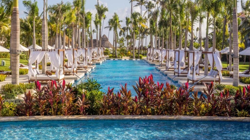 A luxurious pool surrounded by cabanas and palm trees, with manicured gardens and vibrant red plants in the foreground under a clear blue sky.