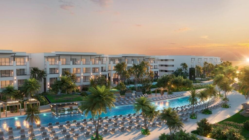 A modern resort with multiple buildings, palm trees, a long pool surrounded by lounge chairs, and a sunset in the background.