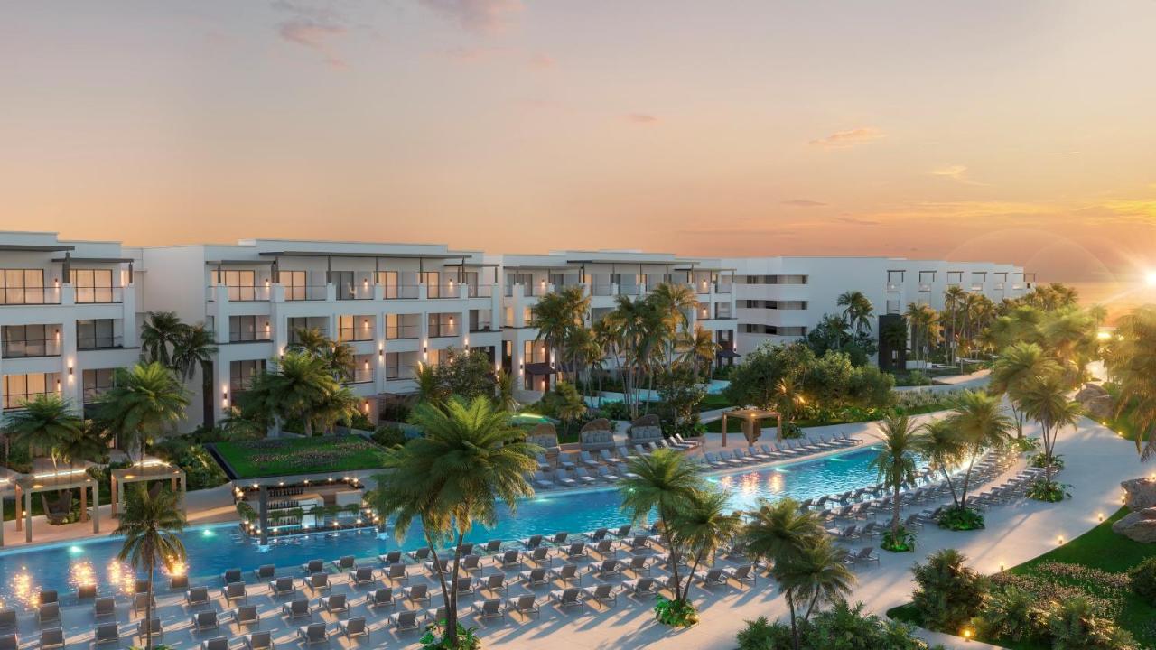 A modern resort with multiple buildings, palm trees, a long pool surrounded by lounge chairs, and a sunset in the background.