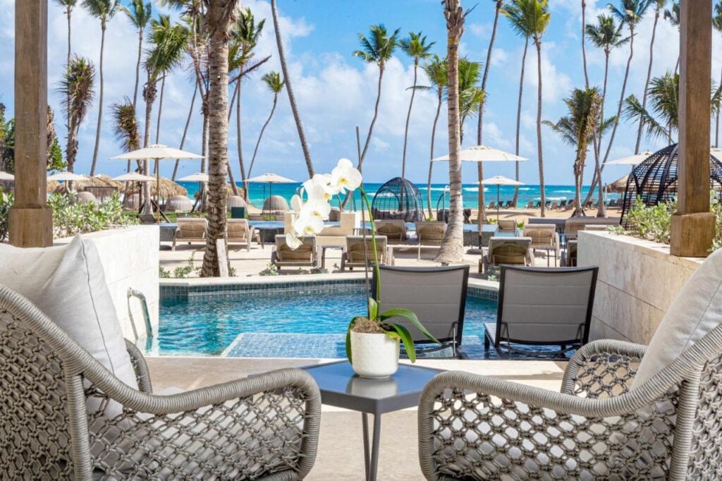 A tropical resort scene featuring a patio with two chairs, a small table with a potted orchid, and a view of a swimming pool, sun loungers, palm trees, and the ocean in the background.