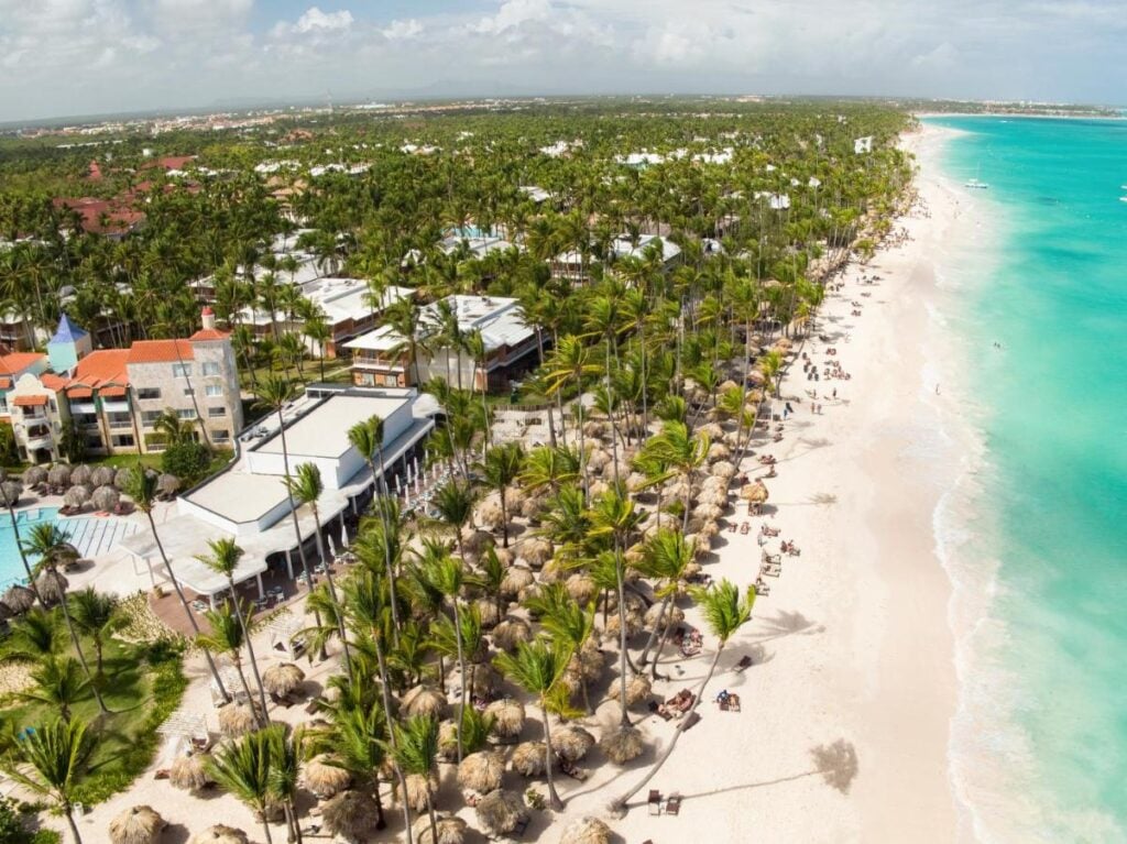 Aerial view of a tropical beachfront lined with palm trees, resort buildings, and white sandy beach. People are seen relaxing on the beach and swimming in the turquoise water.