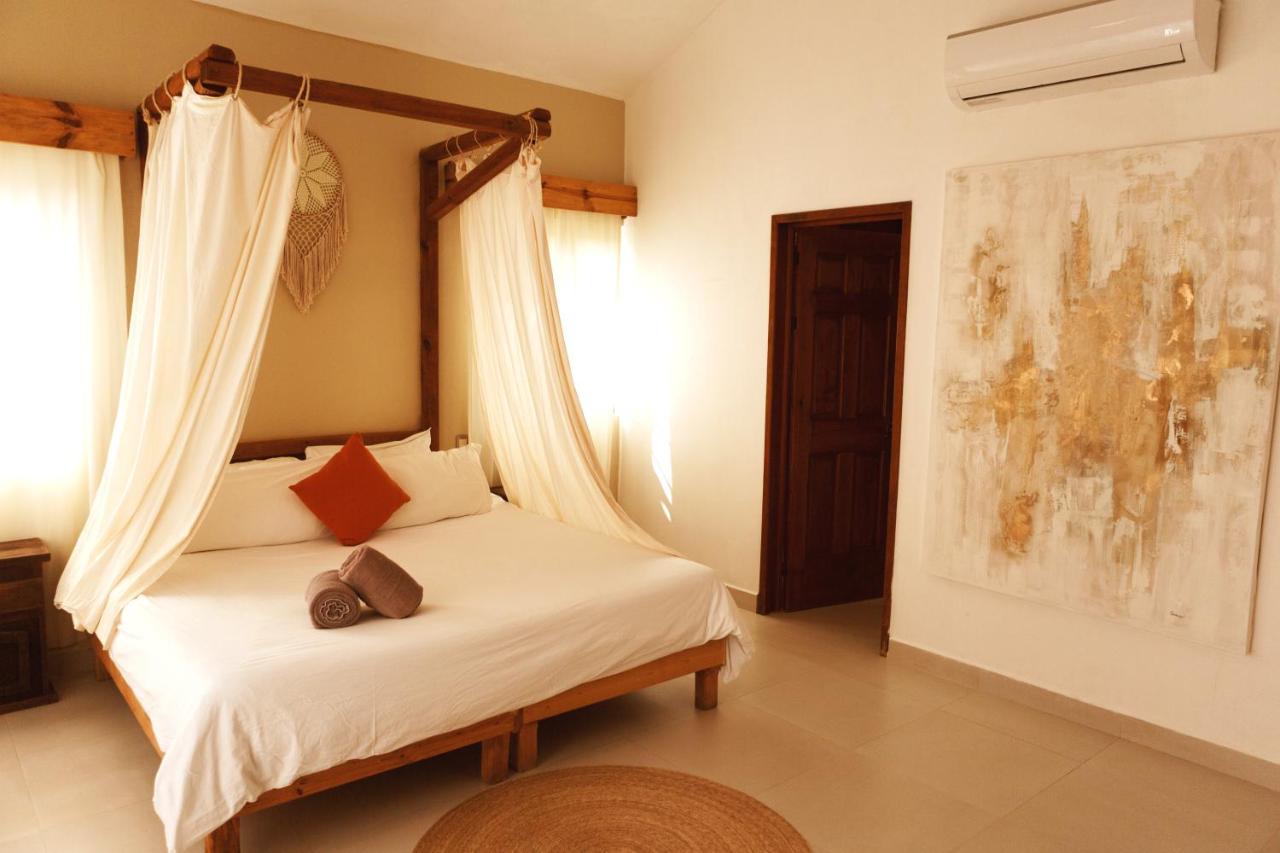 A bedroom with a four-poster bed draped with white curtains, a red cushion, and two rolled towels. The room has beige walls, an abstract painting, AC unit, wooden door, and natural light—a perfect spot if you're wondering where to stay in Cancun.