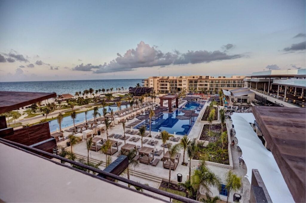 Aerial view of a large resort featuring multiple swimming pools, lounge chairs, and palm trees, with buildings in the background and an ocean view at sunset—truly a stunning option for where to stay in Cancun.