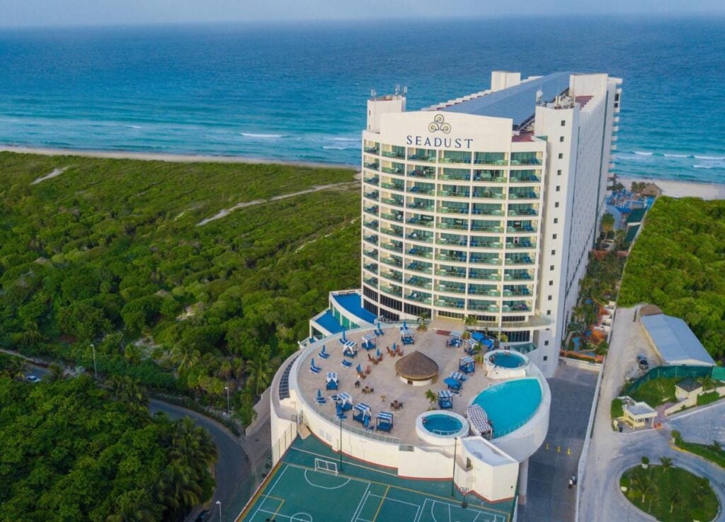 Aerial view of Seadust Cancun Family Resort, a top pick for where to stay in Cancun. The resort features a large building with a rooftop deck, two round pools, a sports court, and a nearby beach surrounded by dense green vegetation.