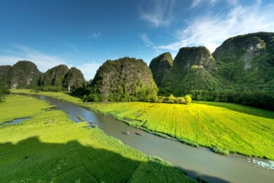 A river winds through lush green fields and karst hills under a clear blue sky.
