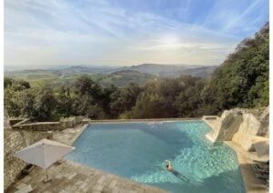 A guest swims in a rectangular outdoor pool surrounded by trees, with a view of green hills and a clear sky at an eco hotel in Italy.