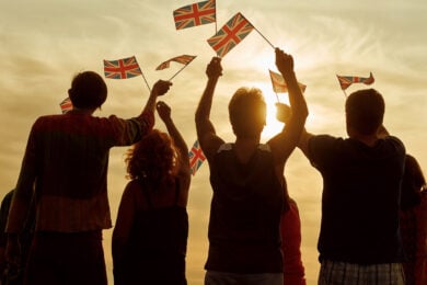 A group of people stand together at sunset, holding and waving several Union Jack flags.