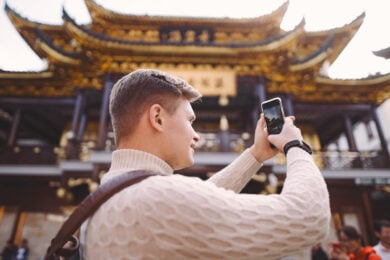 A man in a light sweater and backpack takes a photo with his phone in front of a traditional Asian temple with ornate golden details.