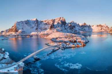 Aerial view of a snow-covered village connected by a bridge, surrounded by calm water and rugged, snow-capped mountains under clear blue sky.