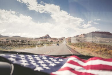 View from inside a car driving down a desert highway, with an American flag draped on the dashboard and rock formations visible ahead under a partly cloudy sky.