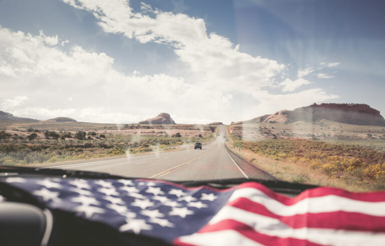 View from inside a car driving down a desert highway, with an American flag draped on the dashboard and rock formations visible ahead under a partly cloudy sky.