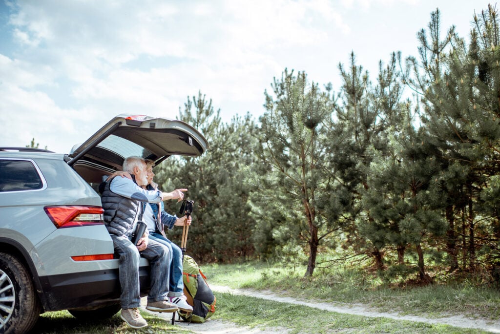 Two older adults sit at the open trunk of an SUV, holding hiking poles and looking toward a pine forest on a sunny day.