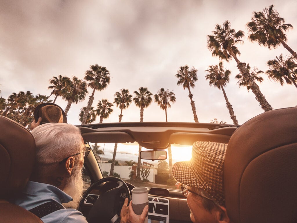 Two people drive in a convertible with the top down, passing palm trees at sunset. One holds a cup, and both wear hats. The sky is partly cloudy.