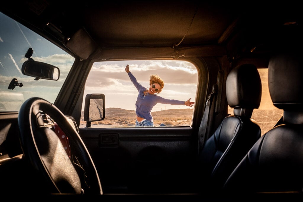 A child wearing sunglasses and a blue shirt jumps with arms wide open outside a parked car in a desert landscape, seen through the car’s open window.