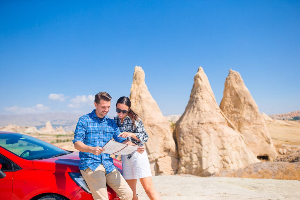 A man and woman stand next to a red car, looking at a map, with unique rock formations and a clear blue sky in the background.