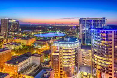 Aerial view of downtown Orlando, Florida, at sunset with illuminated high-rise buildings and a lake in the background.