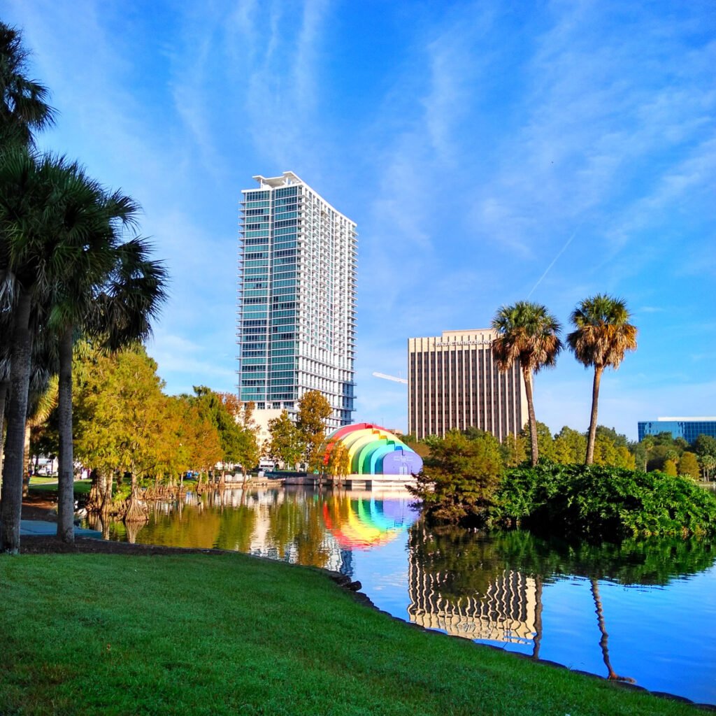 A city park with palm trees and a pond reflecting nearby buildings and a rainbow-colored structure under a blue sky.
