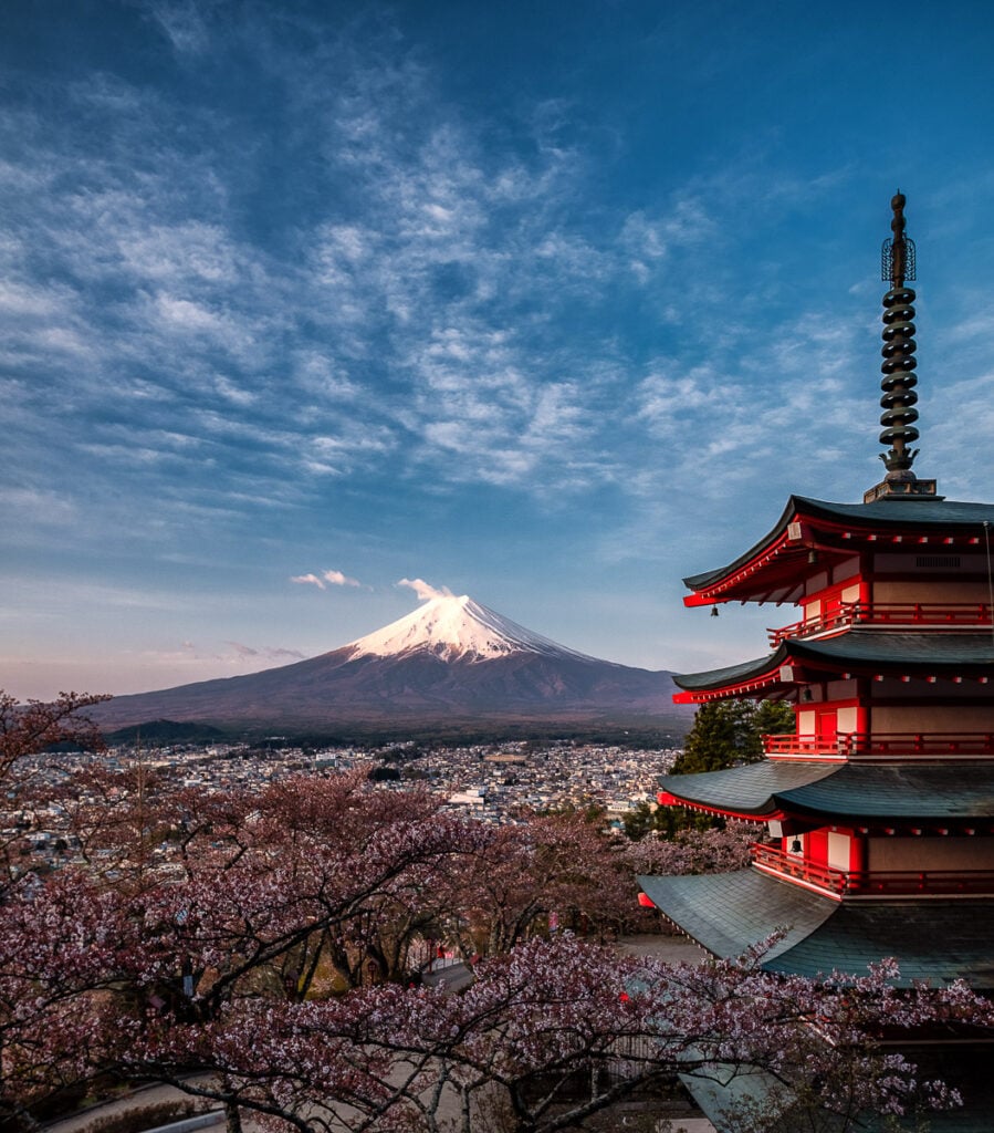 A traditional Japanese pagoda with cherry blossoms in the foreground and snow-capped Mount Fuji in the background under a blue sky.