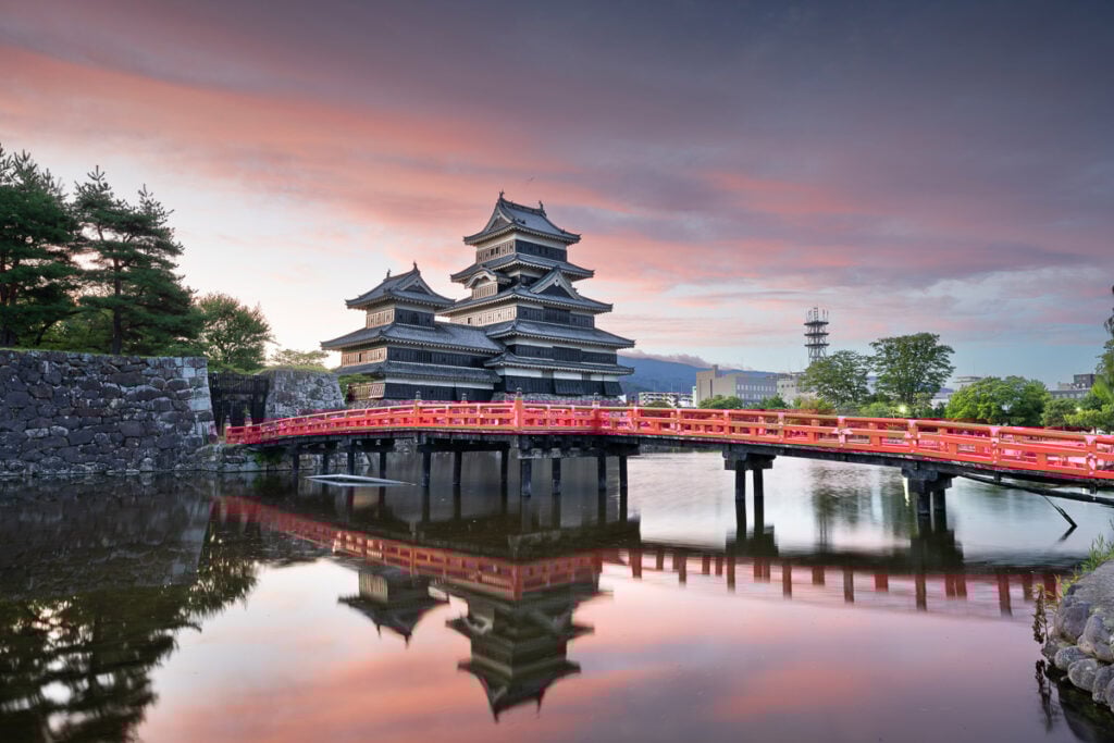 A traditional Japanese castle with a red bridge is reflected in a calm moat at sunset, with trees and a cityscape in the background.