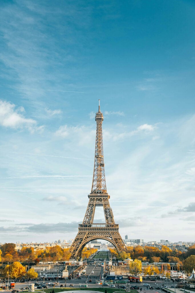 The Eiffel Tower stands in the center of the image under a blue sky, with trees and buildings surrounding its base and people visible in the foreground.