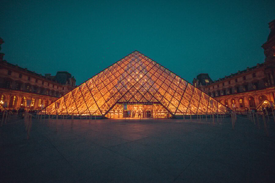 The illuminated glass pyramid entrance of the Louvre Museum in Paris stands prominently at dusk, framed by the museum’s historic building.