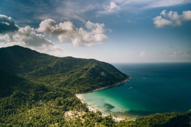 Aerial view of a lush green coastline with a curved beach, turquoise water, and hills under a partly cloudy blue sky.