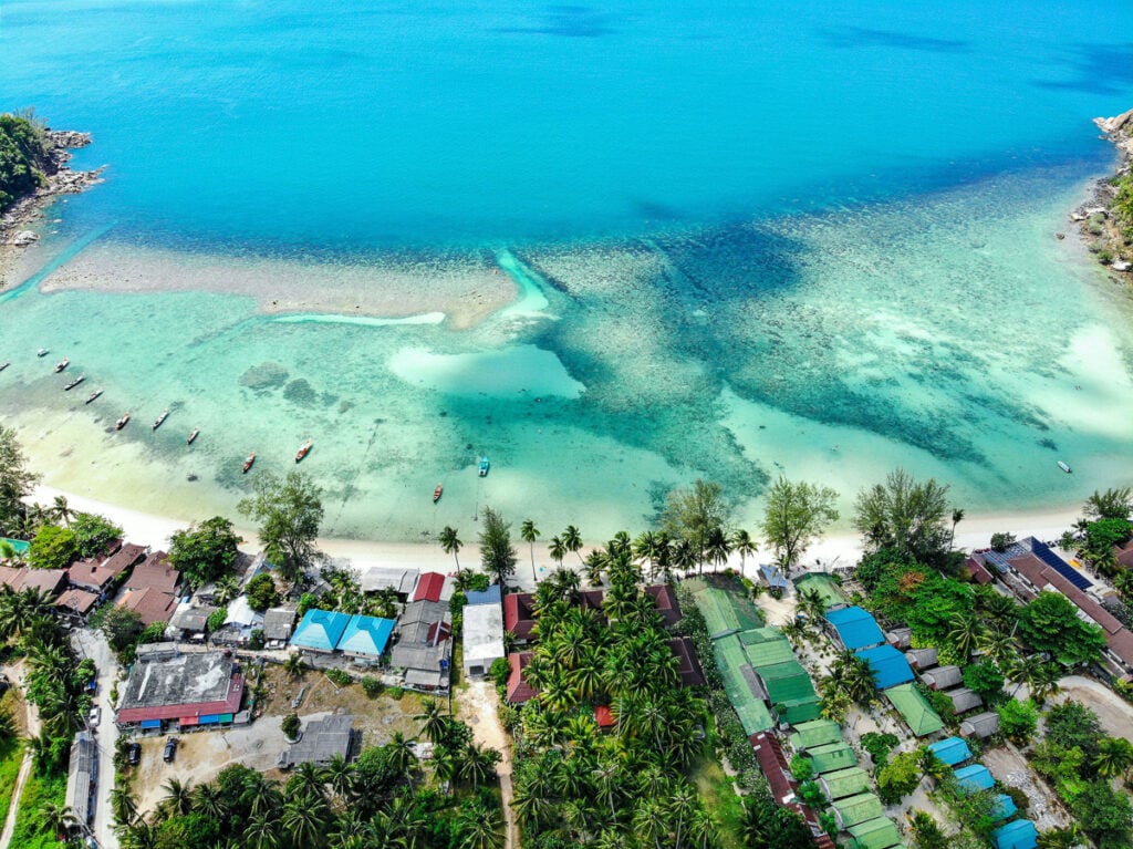 Aerial view of a tropical beach with turquoise water, small boats near the shore, lush palm trees, and buildings lined along the coastline.