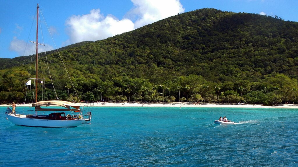 A sailboat anchored near a tropical beach with lush green hills behind, and a small motorboat carrying people on the blue water.
