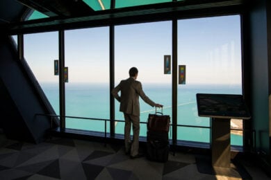A man in a suit stands with luggage, looking out large windows at a view of the sea from a high-rise building.